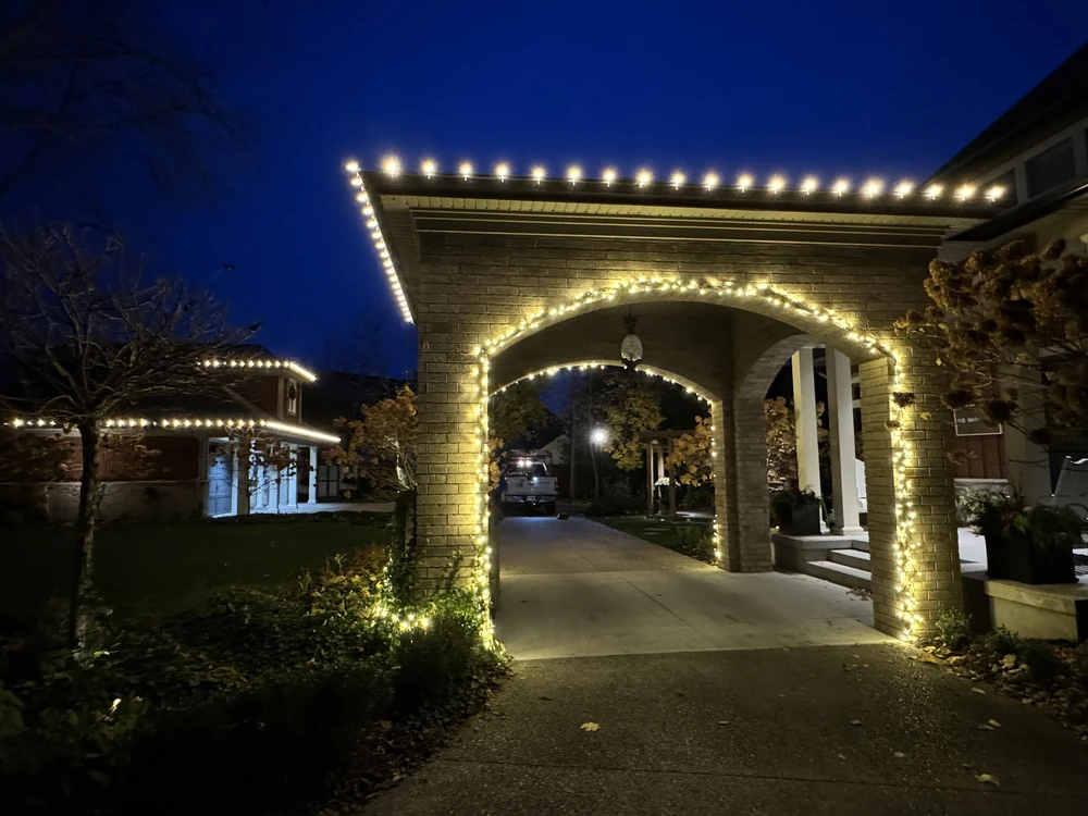 A complete holiday light install showcasing an illuminated brick breezeway, with the work of professional holiday light installers visible in the background.