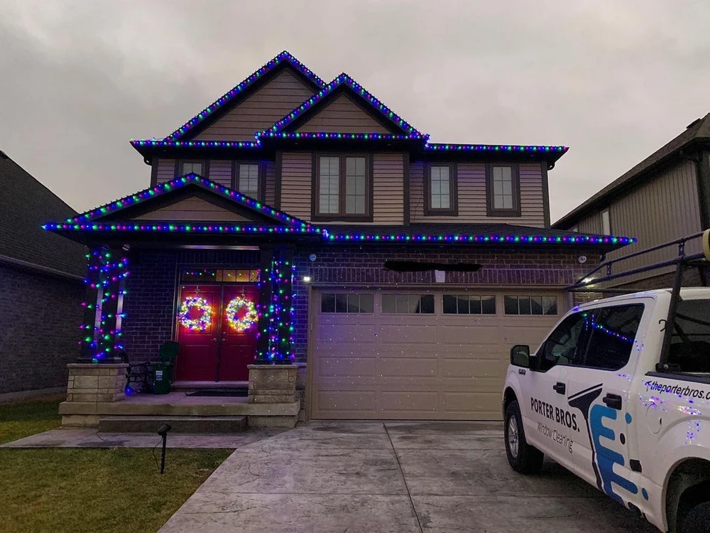 Custom Christmas light decorating on a two-story home using vibrant blue, green, and purple lights along the roof and pillars, with the Porter Bros truck in the driveway.